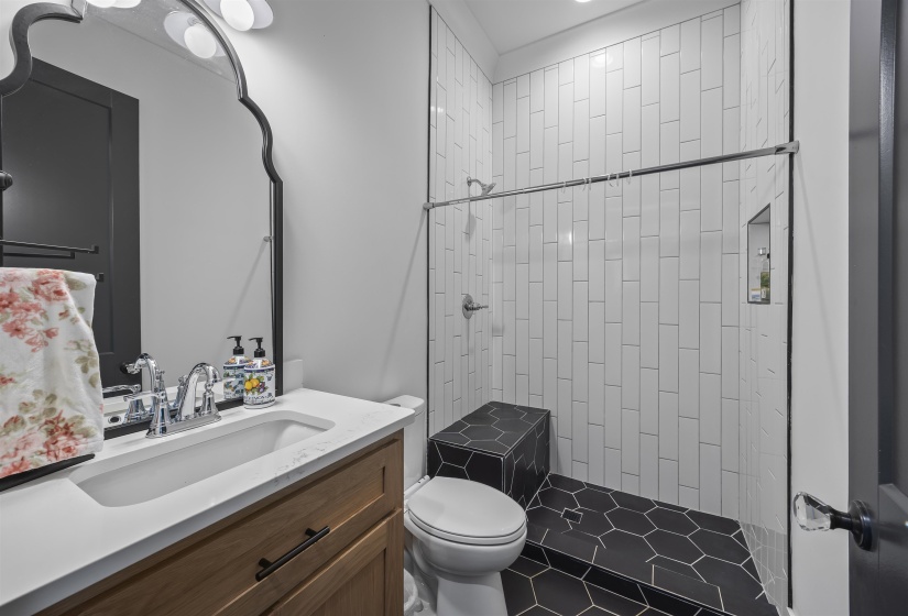 Bathroom featuring a wood-finish vanity with a white countertop and integrated sink, a dark framed mirror, and a walk-in shower with white subway tile walls and black hexagonal tile flooring