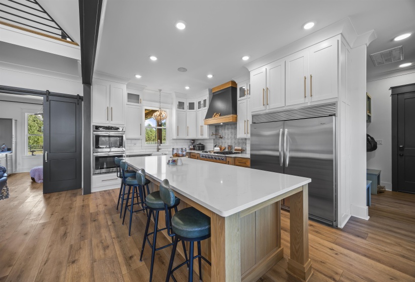 Gourmet kitchen featuring a large island with seating, stainless steel appliances, white cabinetry with gold-tone hardware, wood-finish flooring, and a unique range hood with wood accents
