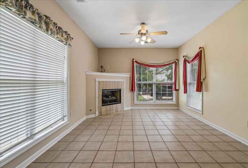 Spacious room featuring neutral paint tones, tiled flooring, a corner fireplace with a light-toned surround, a ceiling fan, and multiple windows with blinds
