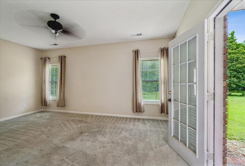 Carpeted interior space featuring a ceiling fan, two windows with blinds and drapes, and a glass-paned door opening to an exterior brick wall and green space