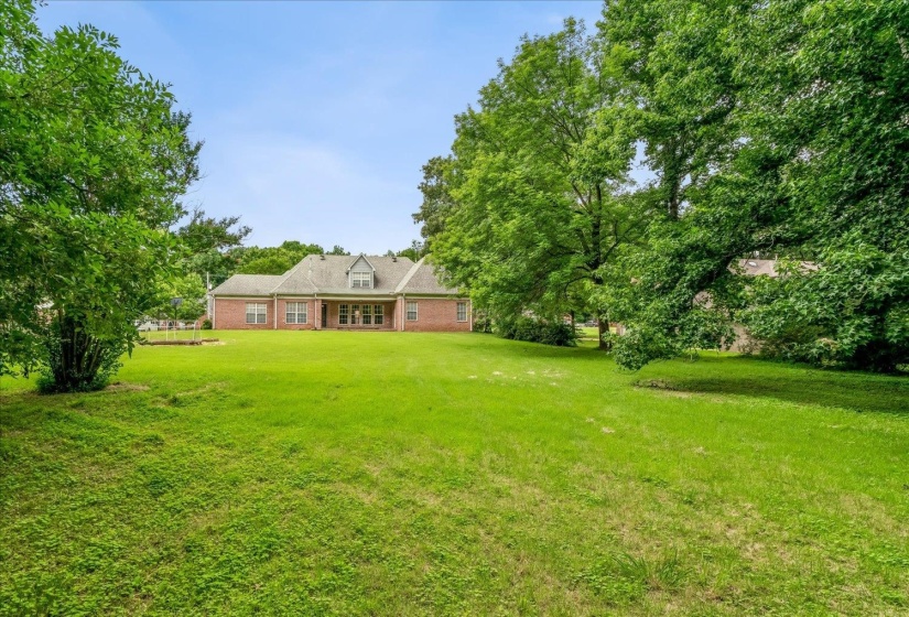 Expansive green lawn with mature trees and a brick exterior residence featuring a dormer window, multiple windows, and a covered patio