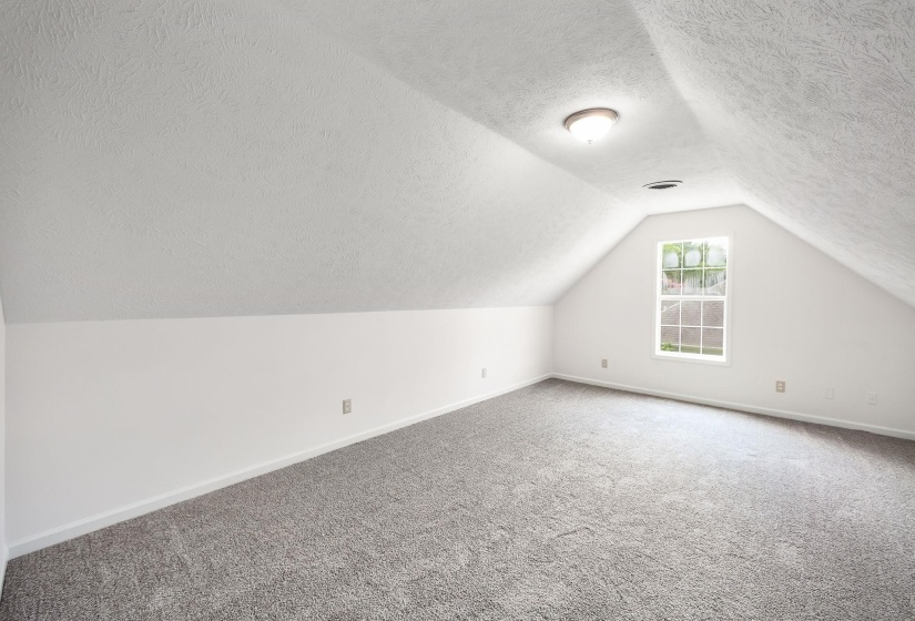 Attic space featuring sloped ceilings, a single window with grid panes, textured white walls, and gray carpet flooring