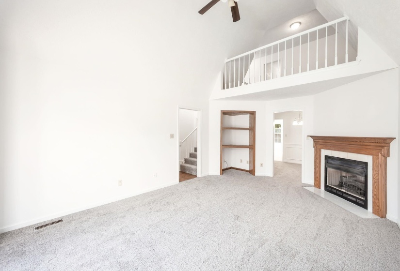 Vaulted living area featuring a wood-mantled fireplace, built-in shelving, a ceiling fan, and a balcony overlook