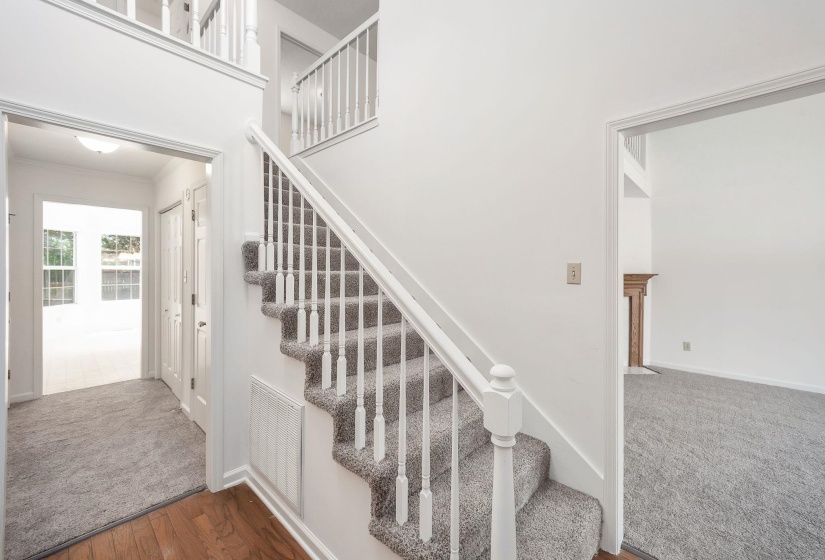 Foyer with wood-finish flooring transitioning to gray carpeting, featuring a white staircase with balusters and a second-story railing, and a coat closet with bi-fold doors