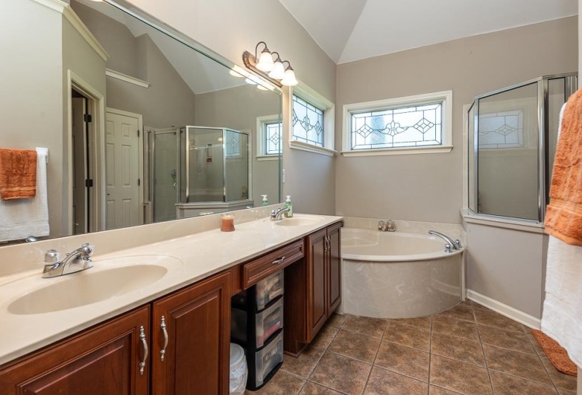 Double vanity with integrated sinks, a built-in corner bathtub, stained-wood cabinetry, ceramic tile flooring, and decorative transom windows