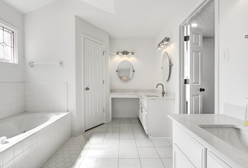 Spacious bathroom featuring a built-in corner tub with tiled surround, a decorative stained-glass window, two separate vanity areas with integrated sinks, and square tiled flooring