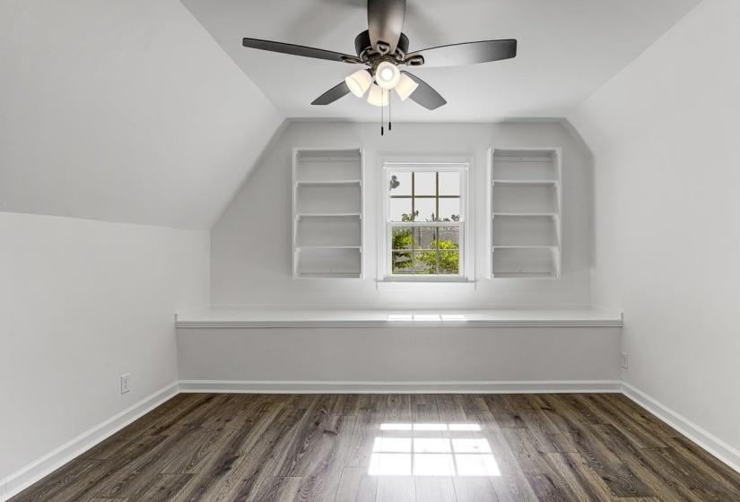 Attic room featuring wood-finish flooring, a window with exterior views, two built-in shelving units, and a ceiling fan with integrated lighting