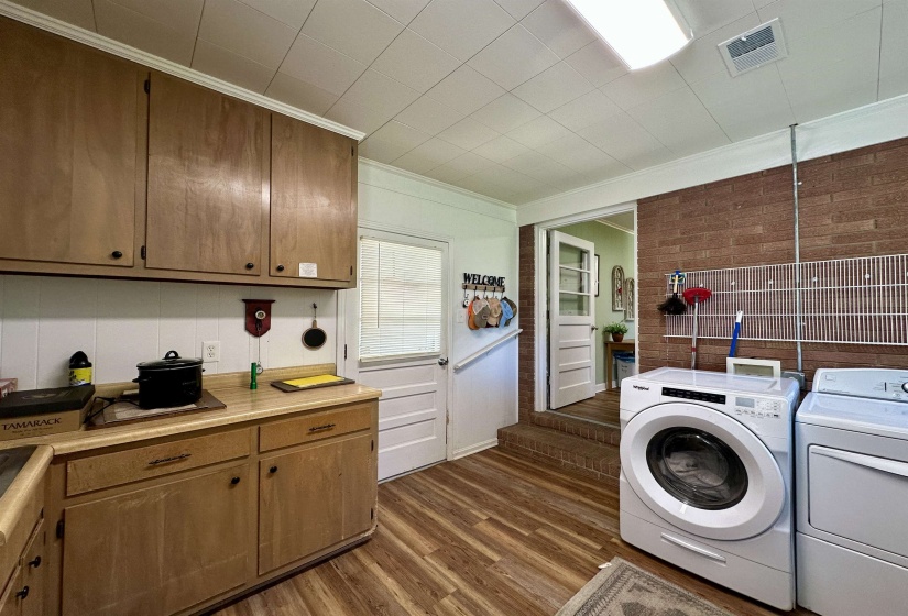 Kitchen and laundry area featuring wood-finish flooring, light wood cabinetry, a white panel backsplash, and a brick-finish accent wall