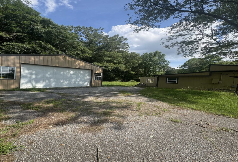 Gravel parking area fronting a garage with light-toned siding and multiple windows, surrounded by mature trees and natural landscaping