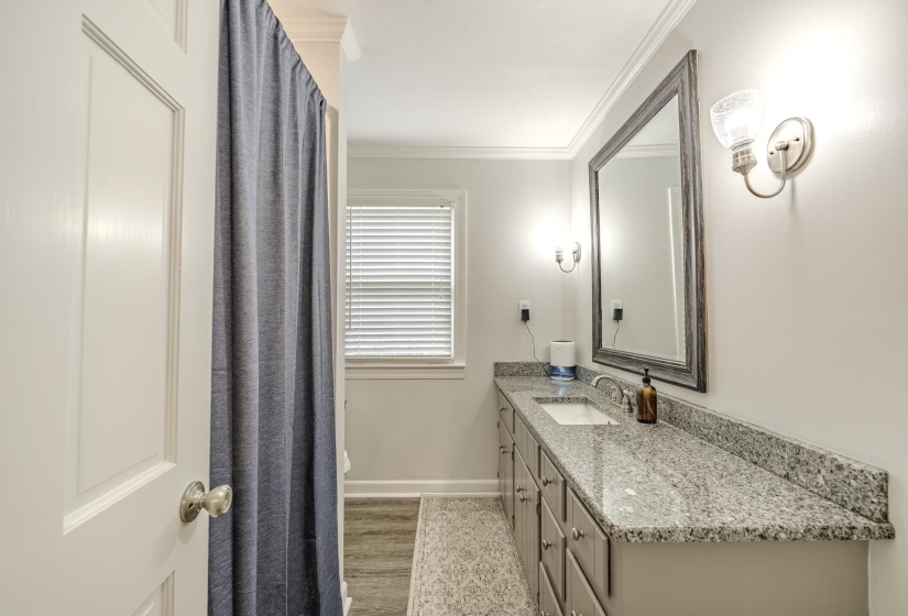 Bathroom featuring a granite-finish countertop, dual wall-mounted light fixtures, a framed mirror, and a window with horizontal blinds