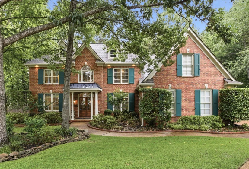 Classic brick exterior featuring a covered entry porch, white window frames with teal shutters, and a well-maintained front lawn with mature trees