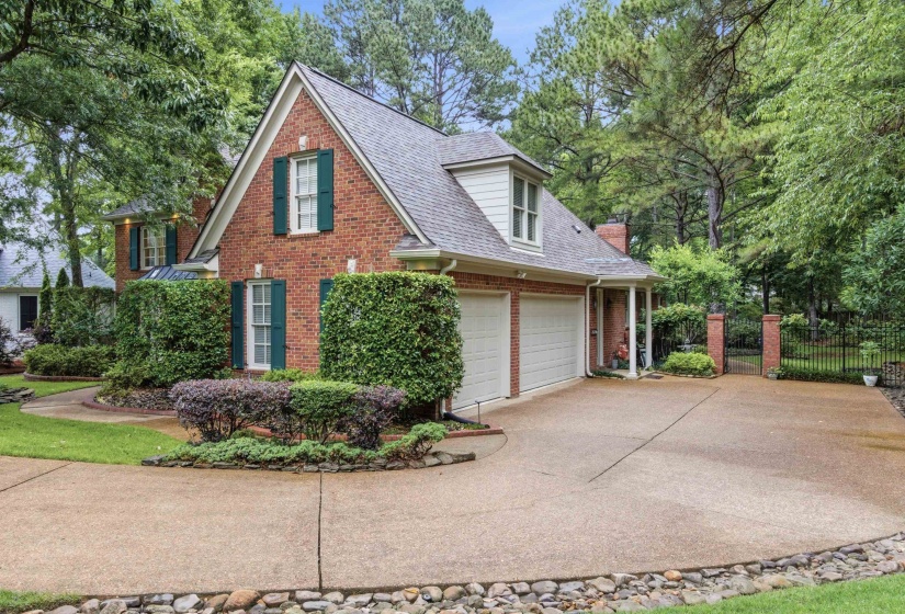 Brick exterior home featuring a gabled roofline, green shutter accents, a two-car garage, mature landscaping, and a paved driveway