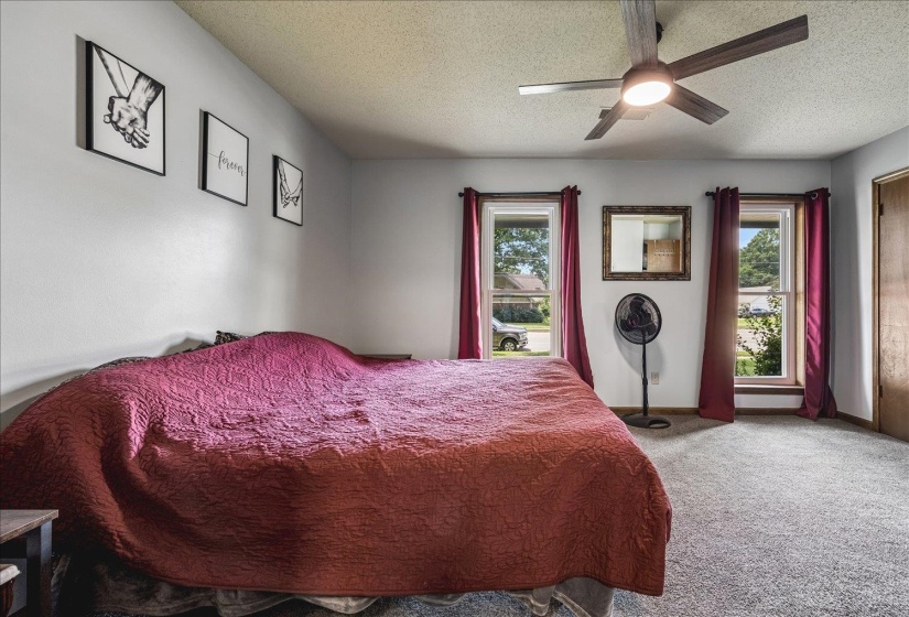 Bedroom featuring two windows providing natural light, a ceiling fan with integrated lighting, textured carpeting, and a light-colored wall finish