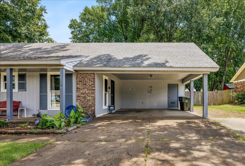 Exterior featuring a carport with an integrated storage door, brick accents, vertical siding, a covered front porch, and mature trees providing shade