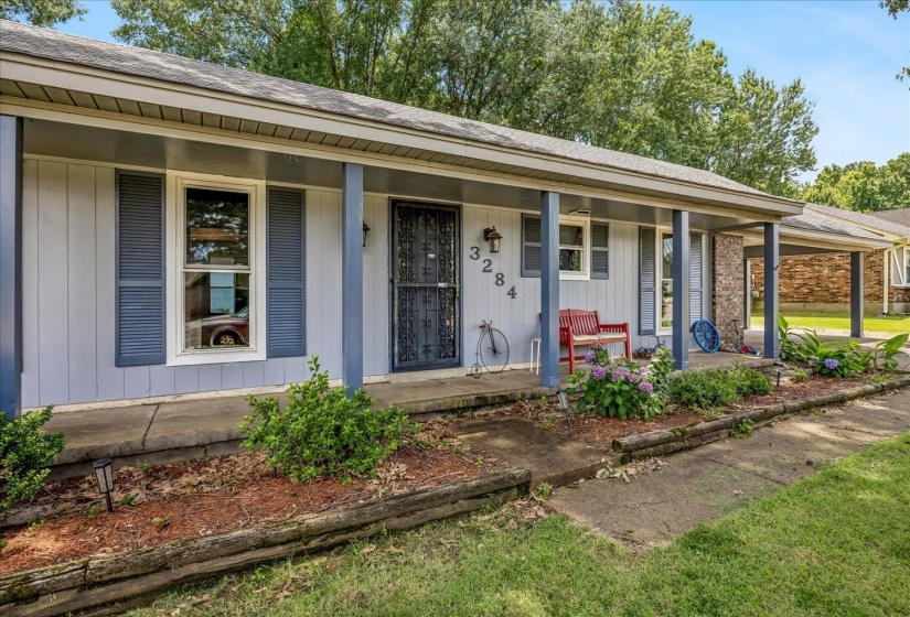 Front exterior featuring light gray siding with blue-gray shutters, a covered porch with supporting columns, and a stone accent wall