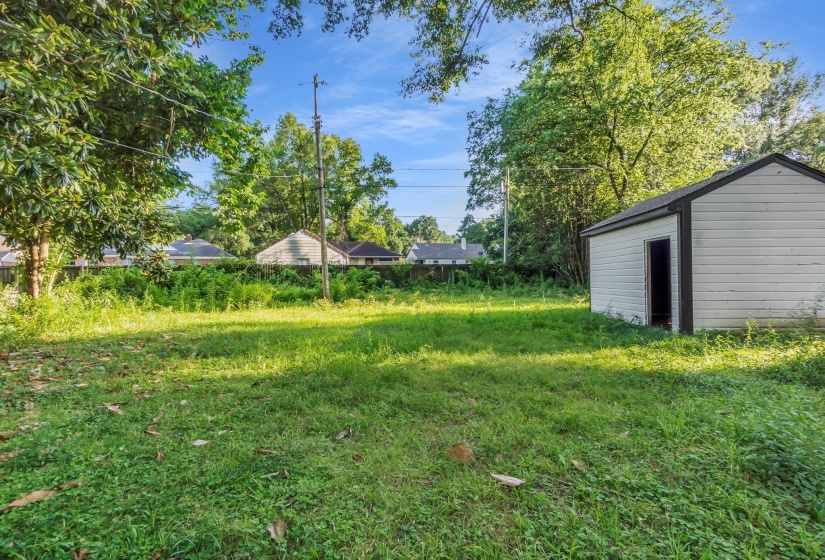 Spacious backyard featuring a detached outbuilding with a single entry, lush green foliage, and natural tree coverage