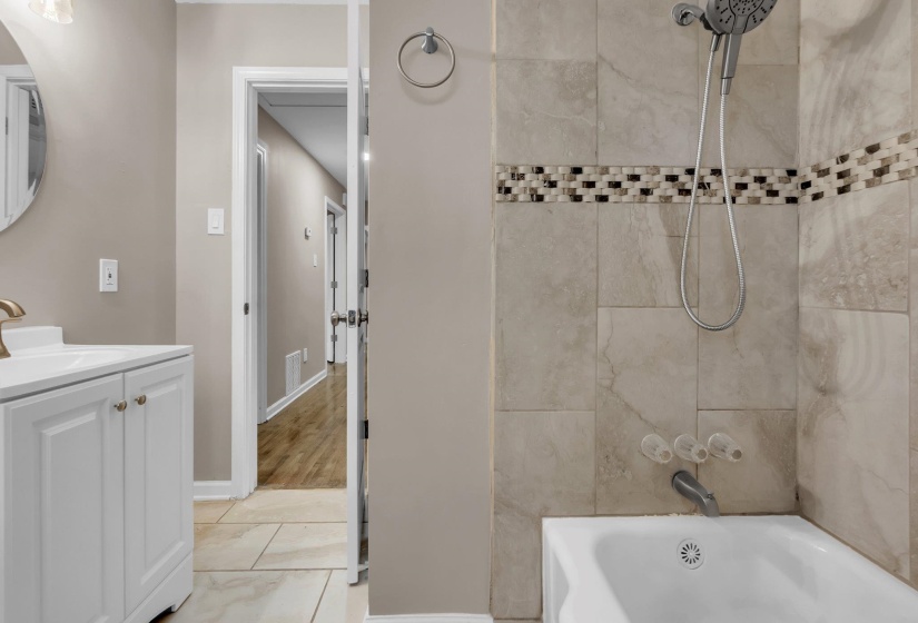 Full bathroom featuring a white vanity with a single basin sink, round mirror, and a tub/shower combination with tile surround and decorative mosaic accent