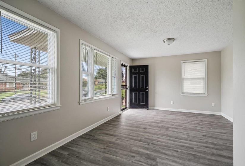 Spacious interior with neutral wall paint, wood-finish flooring, multiple windows with blinds, a prominent black paneled entry door, and a ceiling-mounted light fixture