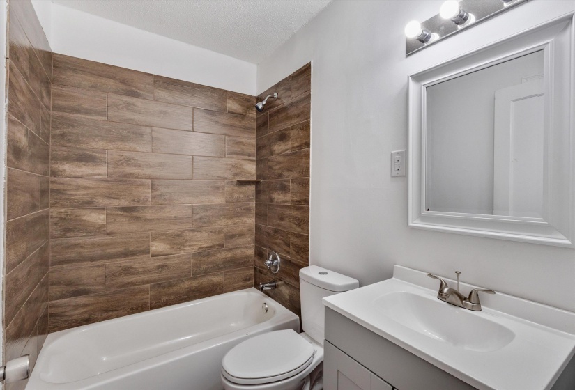Bathroom featuring a white bathtub, wood-finish tile shower surround with integrated shelf, white toilet, and a vanity with a white countertop, undermount sink, and brushed nickel faucet