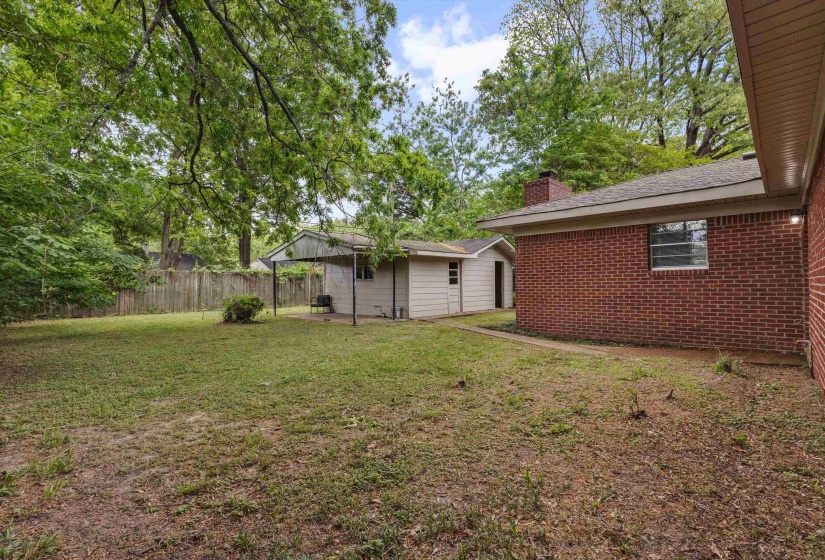 Expansive backyard featuring a brick exterior structure, a separate outbuilding with a covered patio area, and mature trees providing natural canopy