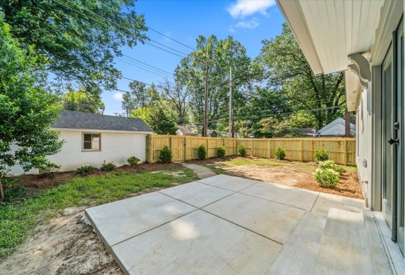 Spacious concrete patio with a newly installed wood privacy fence, featuring a detached structure with a pitched roof, surrounded by mature trees