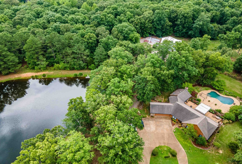 Expansive property featuring a private pond with a small dock, a custom-shaped swimming pool with surrounding patio, a residential structure with a gray shingle roof, and a paved driveway