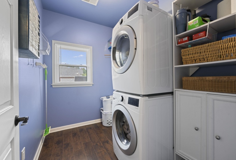 Dedicated laundry area featuring stacked front-load washer and dryer, wood-finish flooring, white cabinetry, open shelving, and a window for natural light