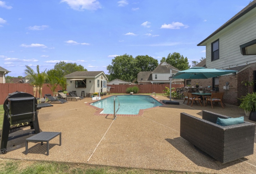 Expansive backyard featuring a custom-shaped swimming pool with concrete decking, a dedicated pool house, a brick patio, and a privacy fence