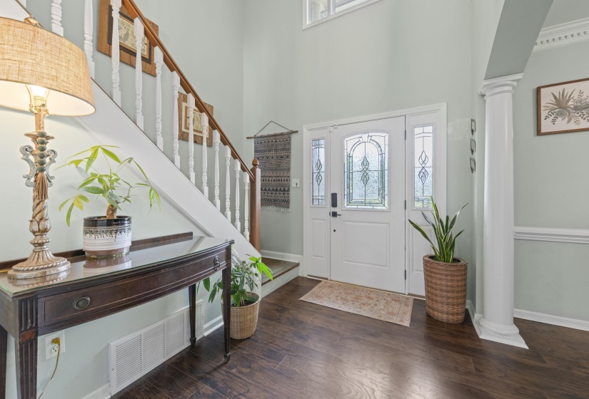 Two-story foyer featuring dark wood-finish flooring, a white front door with decorative glass inserts and sidelights, a prominent staircase with white balusters and a wood handrail, a decorative column, and crown molding