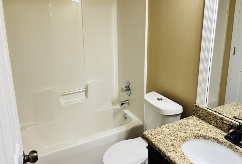 Bathroom featuring a white integrated tub and shower unit, a toilet, and a vanity with a speckled countertop and an undermount sink
