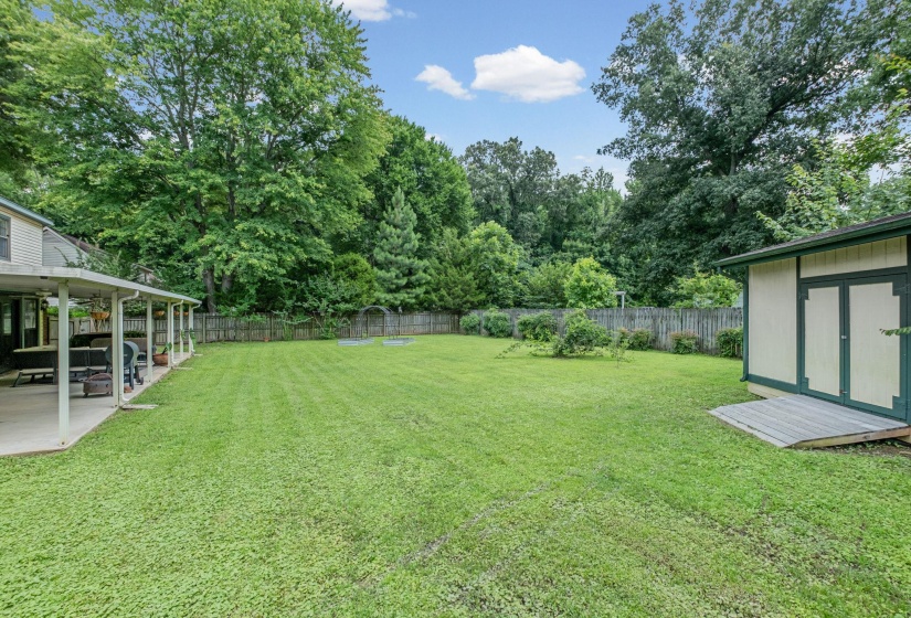 Expansive green lawn featuring a covered patio, a storage shed with a ramp, and a perimeter fence, bordered by mature trees