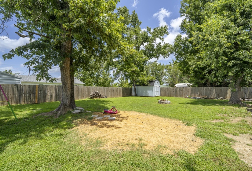 Spacious backyard featuring a privacy fence, mature trees, a storage shed, and a fire pit
