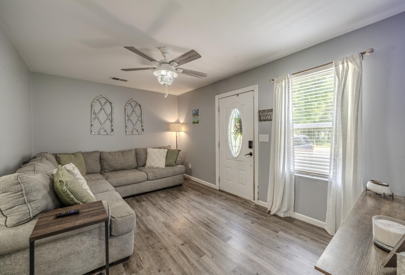 Living space featuring wood-finish flooring, a white front door with an oval leaded glass insert, a window with blinds and white drapes, a ceiling fan with light fixture, and light gray wall paint