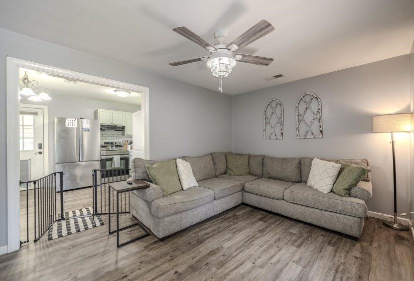 Living area with wood-finish flooring, a ceiling fan with light fixture, and a wide entryway to the kitchen featuring stainless steel appliances