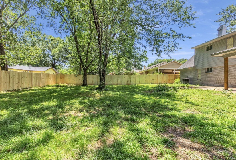 Expansive grassy yard featuring a newly installed privacy fence, mature trees, and a covered patio area with a wood support column