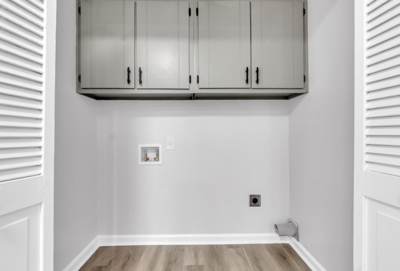 Dedicated laundry alcove featuring overhead cabinetry with black hardware, wood-finish flooring, white baseboards, and louvered bifold doors