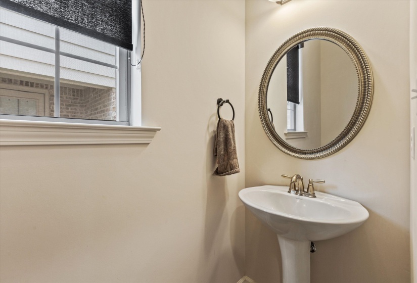 Pedestal sink with a brushed nickel faucet, framed round mirror, and a window featuring a roller shade and white sills