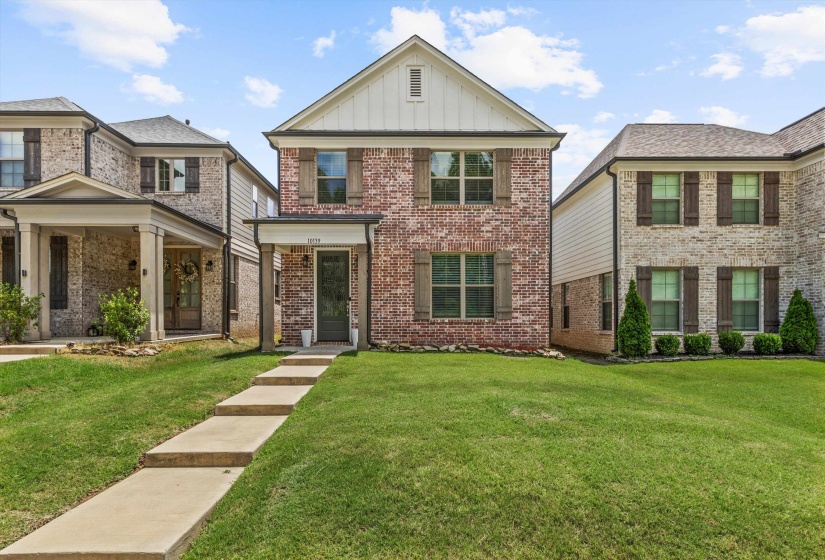 Two-story brick facade featuring a covered entry porch, dark-paned windows with decorative shutters, and a front yard with a concrete walkway