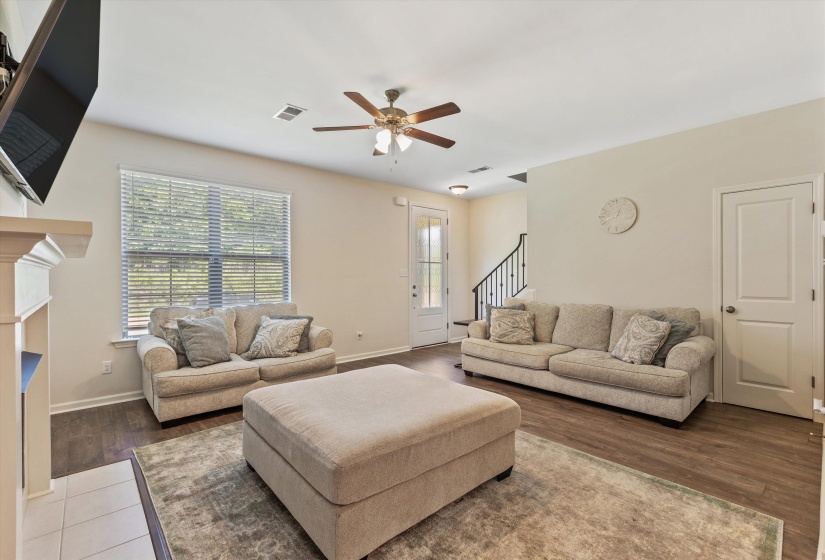 Spacious living area featuring wood-finish flooring, a ceiling fan with integrated lighting, a substantial window with blinds, a white paneled interior door, and a staircase with dark wrought iron spindles
