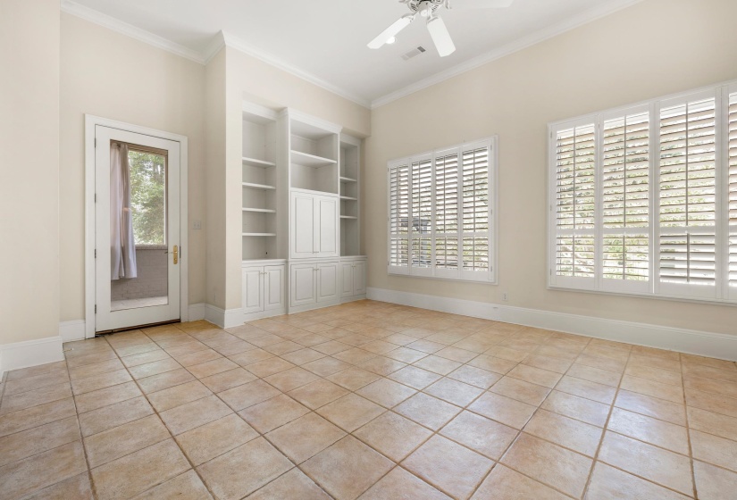 Spacious room featuring neutral-toned tile flooring, extensive built-in shelving with cabinetry, two large windows with plantation shutters, a glass-paneled door, and a ceiling fan