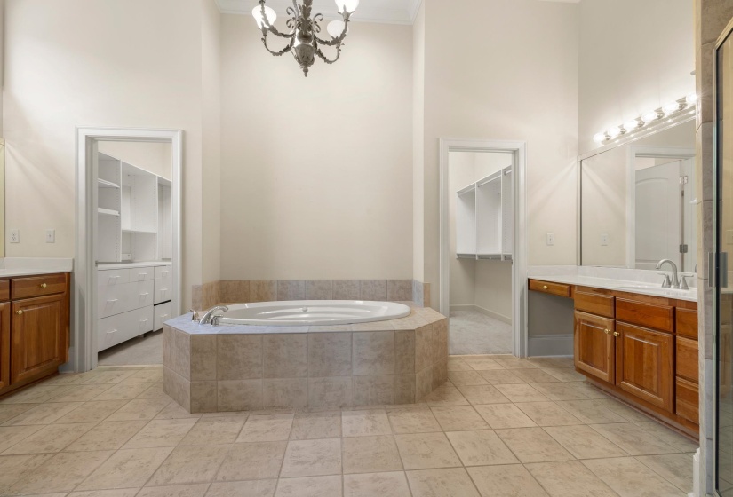 Tiled hexagonal soaking tub with tile surround, dual wood cabinetry vanities with solid surface countertops, and two separate walk-in closet entries