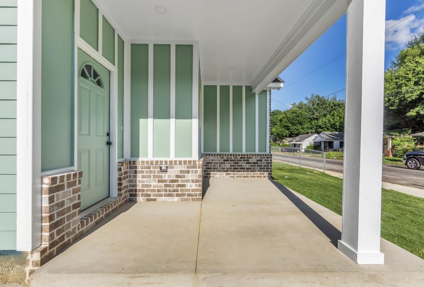 Covered front porch featuring a light green paneled door with a half-moon transom, framed by brick siding and light green vertical siding with white trim