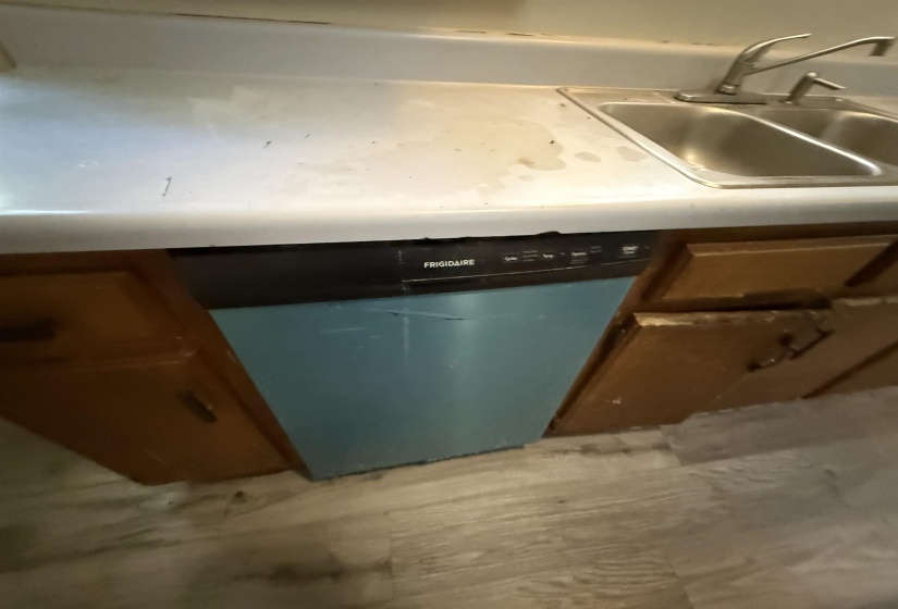 Kitchen area featuring a double basin stainless steel sink with a chrome faucet, a built-in dishwasher with a black control panel, wood-finish base cabinetry, a light-colored countertop, and wood-finish flooring