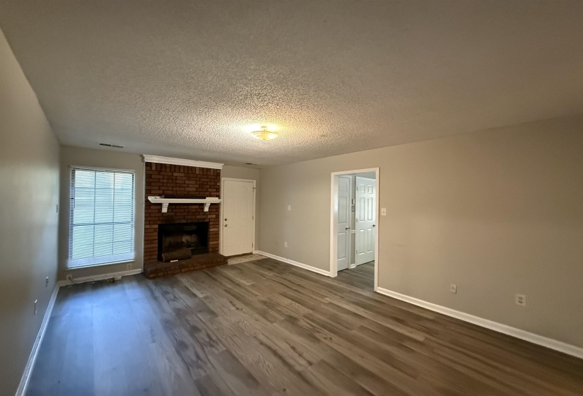 Spacious room featuring wood-finish flooring, a brick fireplace with a white mantel, and neutral wall tones