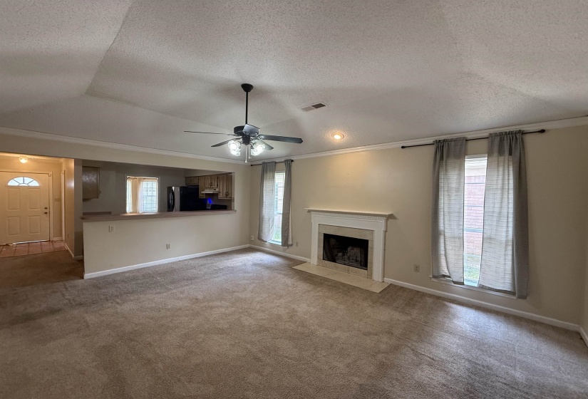 Spacious carpeted living area featuring a central ceiling fan with integrated lighting, a fireplace with a white mantel and hearth, and crown molding