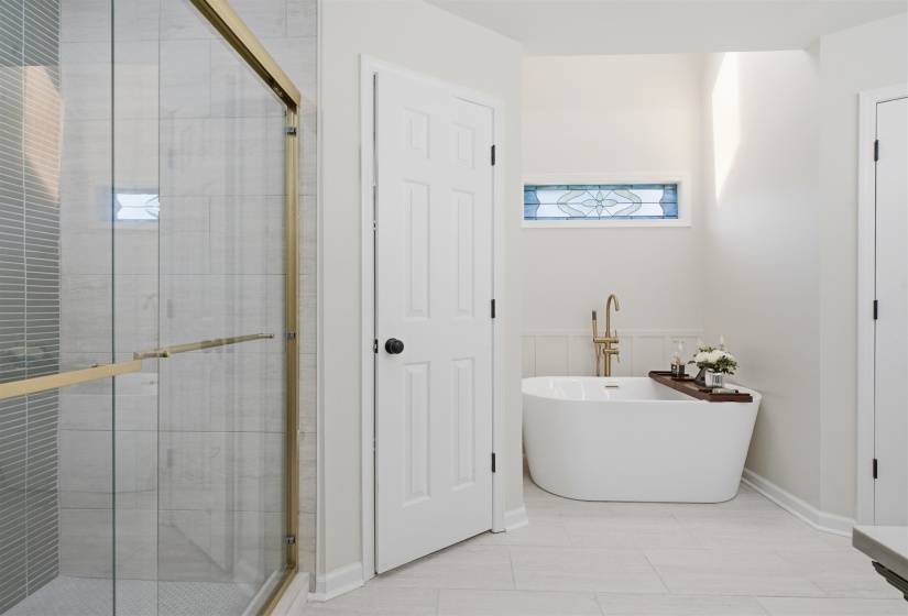Modern bathroom featuring a freestanding soaking tub with a brass-finish floor-mounted faucet, a stained-glass transom window, and a glass-enclosed shower with brass-finish hardware