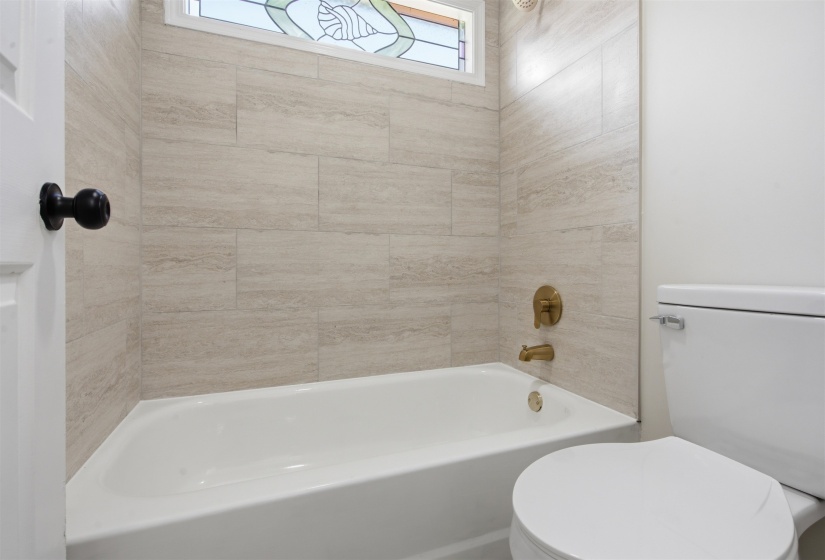 Tub and shower combination featuring beige stone-look tile surround, brushed gold fixtures, and a decorative stained-glass window