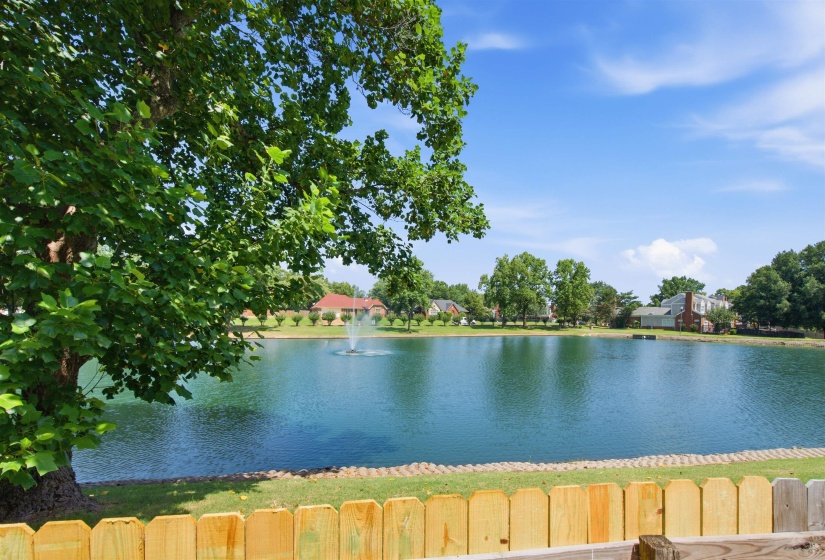 Waterside property featuring a wooden fence, expansive body of water with a fountain, and mature trees framing the perimeter