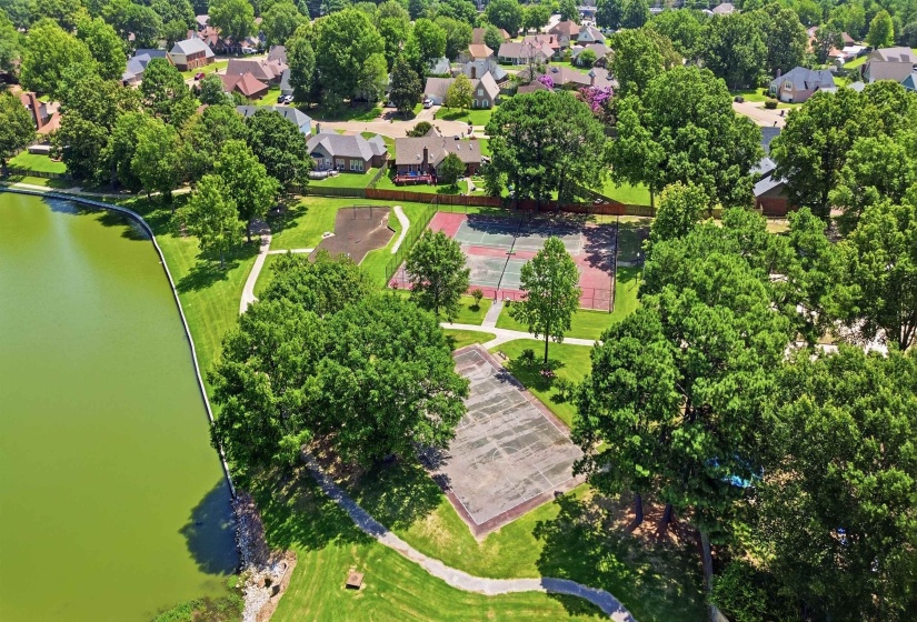 Expansive green space featuring two tennis courts, paved walking paths, and mature trees bordering a large body of water