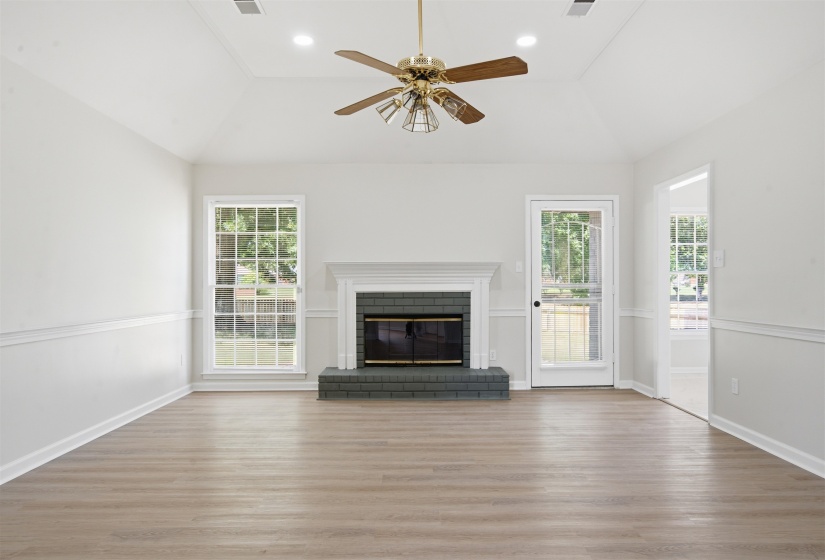 Bright living space featuring a vaulted ceiling with a brass-finish ceiling fan, wood-finish flooring, and a fireplace with a white mantel and dark brick surround